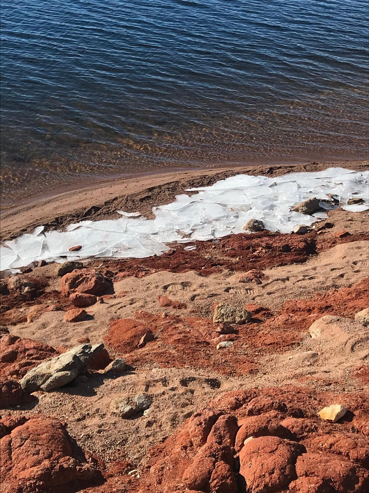 Rivage vu de haut : l’eau sombre, une bande de plaques de glace échouées, la roche rouge.